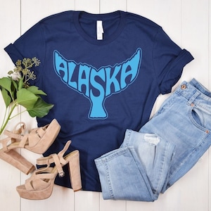 May include: A navy blue t-shirt featuring a blue whale tail graphic that spells out "ALASKA". The shirt is styled with light blue jeans and beige platform sandals.