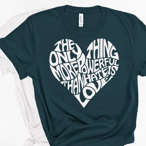 May include: A dark teal short-sleeved t-shirt featuring a large white heart graphic on the chest. The heart is filled with the text, THE ONLY THING MORE POWERFUL THAN HATE IS LOVE. A plain white t-shirt is partially visible underneath the dark teal shirt.