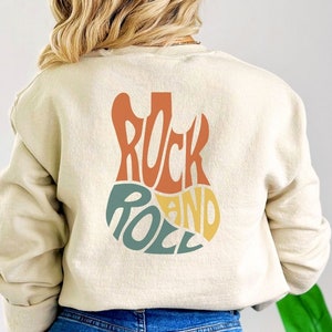 May include: A cream colored sweatshirt with a graphic of a guitar on the back. The guitar is made up of the words "Rock and Roll" in a retro font with orange, yellow, green, and blue colors.