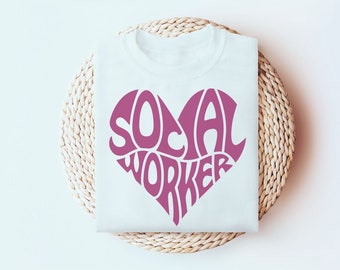 Puede incluir: Una camiseta blanca con un gráfico grande de color rosa oscuro en forma de corazón. El texto "SOCIAL WORKER" está impreso dentro de la forma del corazón en una fuente audaz, estilizada y ondulada que llena toda el área del corazón de arriba a abajo.