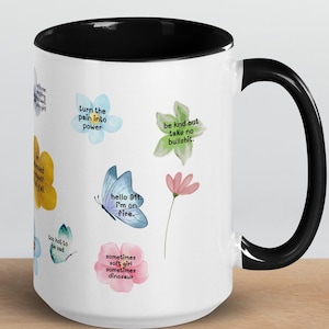 Può includere: Una tazza in ceramica bianca con interno e manico neri, decorata con colorati motivi floreali e a farfalla. La tazza presenta citazioni ispiratrici come "turn the pain into power" e "hello ft I'm on fire."