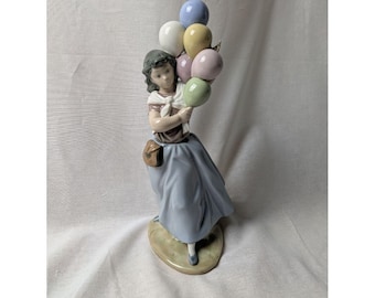 Könnte beinhalten: Porzellanfigur eines Mädchens, das einen Strauß bunter Luftballons hält. Das Mädchen trägt einen blauen Rock, ein braunes Oberteil und eine kleine Tasche. Die Ballons sind in Blau-, Gelb-, Rosa- und Grüntönen gehalten. Die Figur steht auf einem hellgrünen ovalen Sockel.