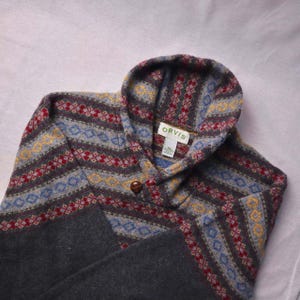 May include: A patterned sweater in shades of grey, blue, red, and yellow, featuring a shawl collar and a single brown button. The label reads "ORVIS".