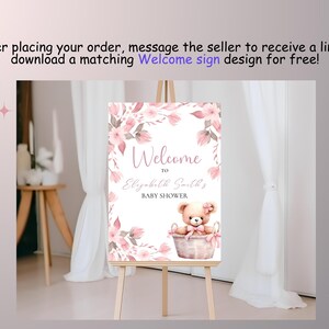 May include: A pink and white welcome sign with floral designs and a teddy bear in a basket. The sign reads "Welcome to Elizabeth Smiths Baby Shower".