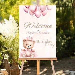 Op de afbeelding: Welkom op Sophia Smith's verjaardagsfeest bord met een roze en witte aquarel achtergrond. Een teddybeer met een roze tutu en een kroon houdt een roze ballon vast. Het bord staat op een ezel.