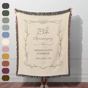 May include: A cream-colored woven throw blanket with fringed edges, featuring a dark brown line-art botanical frame. The text reads 25th Anniversary, Margaret & Peter Richardson, and Since June 27, 1999, with a small heart icon centered.