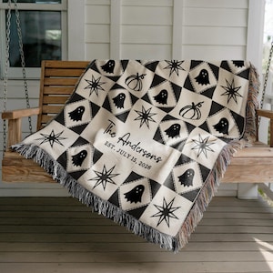 May include: A cream-colored Halloween throw blanket with a repeating pattern of black ghosts, stars, and pumpkins. The blanket is personalized with the text "The Andersons" and the date "EST. JULY 15, 2026". The blanket has a fringed edge.