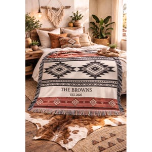 May include: A cream-colored woven blanket with a geometric black and rust-colored pattern. The blanket has a fringe and the text "THE BROWNS EST. 2026". The blanket is on a bed with decorative pillows and a cowhide rug.