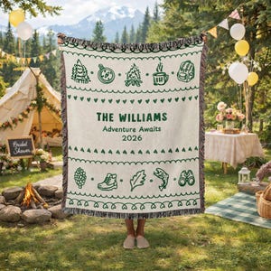 May include: A cream-colored woven blanket with green camping-themed illustrations and the text "THE WILLIAMS Adventure Awaits 2026." The blanket has a fringed edge and is held outdoors.