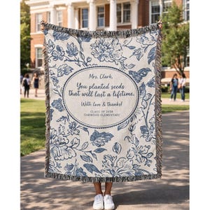 May include: A beige and blue floral throw blanket with a fringed edge. The blanket features a floral pattern and an oval design with text that reads "Mrs. Clark, You planted seeds that will last a lifetime. With love & thanks! CLASS OF 2026 OAKWOOD ELEMENTARY."