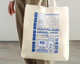 May include: Cream-colored tote bag with blue graphics and text. The design includes a globe, envelopes, a computer, and the phrase "YOU'VE GOT MAIL." Additional text is in multiple languages. The bag is held by a person.