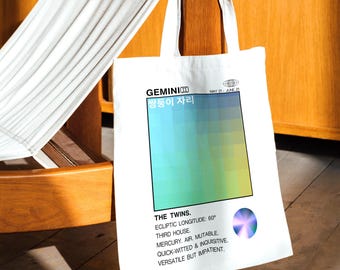 May include: White tote bag with a Gemini zodiac design. The bag features a color gradient and text including "GEMINI", "THE TWINS", and astrological details. The bag is hanging on a wooden chair.