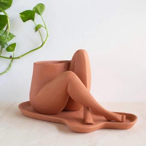 May include: Terracotta planter shaped like a pair of legs sitting on a matching tray. The planter is a unique and modern design for indoor plants.
