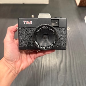 Puede incluir: Cámara negra con la palabra "TIME" en rojo. La lente está etiquetada como "COLOR OPTICAL LENS" y "1:6 f=50mm". La cámara tiene un agarre texturizado. La palabra "Matrix" es visible en el fondo.