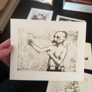 May include: A black and white etching print of a man in a boxing pose. The man is shirtless with a mustache and is wearing black shorts. The print is on a white rectangular paper.