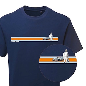 Puede incluir: Camiseta azul marino con un diseño horizontal con rayas naranjas y blancas, un coche de carreras plateado y una figura con un traje de carreras blanco. La palabra "TrackLine" está impresa en blanco. Una incrustación circular muestra un primer plano del diseño.