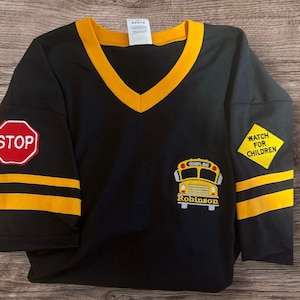 Puede incluir: Camiseta negra de manga larga con rayas amarillas y cuello en V amarillo. La camiseta tiene un autobús escolar bordado con el texto "School Bus" y "Robinson". La manga izquierda tiene una señal de stop roja y la manga derecha tiene un diamante amarillo con el texto "Atención a los niños".