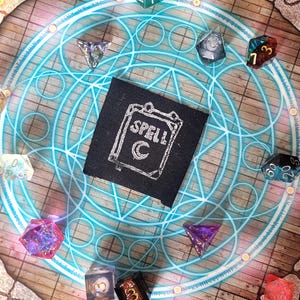 May include: A black fabric patch with a white outline of a book with the word "SPELL" written on it. The book has a crescent moon symbol on the cover. The patch is surrounded by colorful dice on a wooden surface with a blue and white circular pattern.