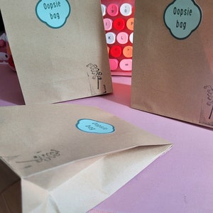 May include: Three brown paper bags with a blue and white sticker that says "Oopsie bag" on each. The bags are on a pink surface.