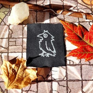 May include: A black fabric patch with a white embroidered design of a bird with a single eye. The bird is standing on a branch.