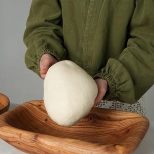 May include: A person in a green jacket is holding a large ball of white dough over a wooden bowl. The bowl is rectangular and has a natural wood finish.