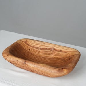 May include: A rectangular wooden serving bowl with a natural wood finish. The bowl has a smooth, rounded interior and a slightly rough exterior. The grain of the wood is visible throughout the bowl.