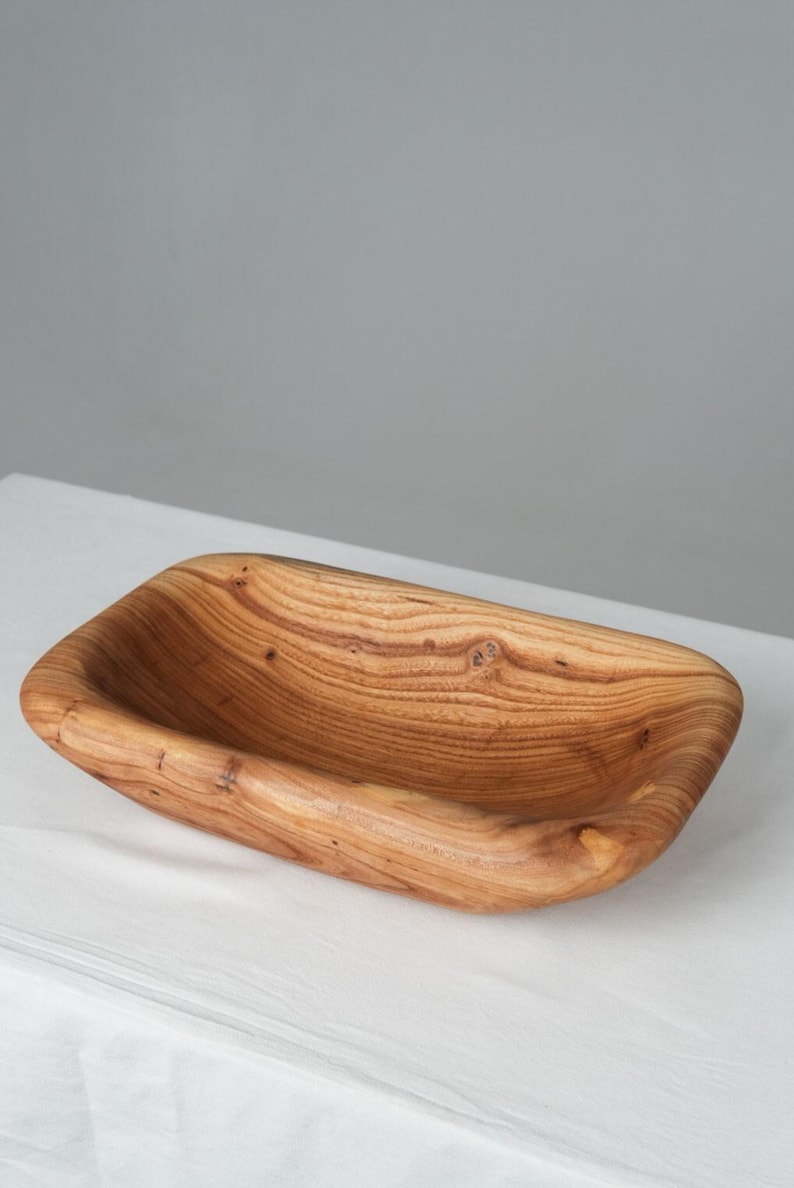 May include: A rectangular wooden serving bowl with a natural wood finish. The bowl has a smooth, rounded interior and a slightly rough exterior. The grain of the wood is visible throughout the bowl.