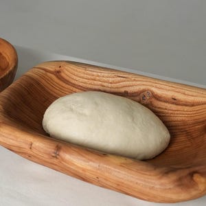 May include: A close-up of a white dough ball resting in a wooden bowl. The bowl is made of light brown wood with a natural grain pattern.