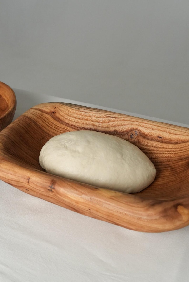 May include: A close-up of a white dough ball resting in a wooden bowl. The bowl is made of light brown wood with a natural grain pattern.