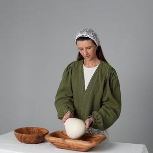 May include: A woman wearing a white lace head covering and a green jacket is placing a ball of dough into a wooden bowl. The bowl is on a white tablecloth.