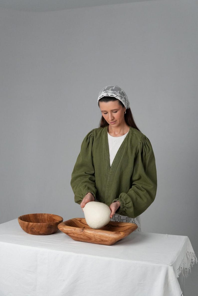 May include: A woman wearing a white lace head covering and a green jacket is placing a ball of dough into a wooden bowl. The bowl is on a white tablecloth.