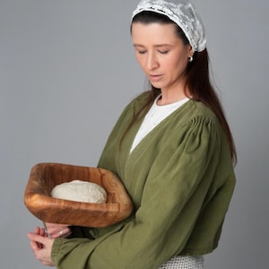 May include: A woman wearing a white lace head covering and a green long-sleeved shirt holds a wooden bowl with a round loaf of dough inside.