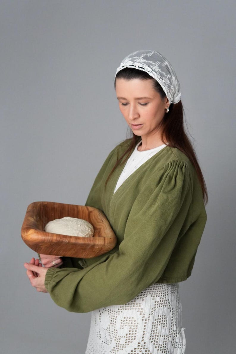 May include: A woman wearing a white lace head covering and a green long-sleeved shirt holds a wooden bowl with a round loaf of dough inside.