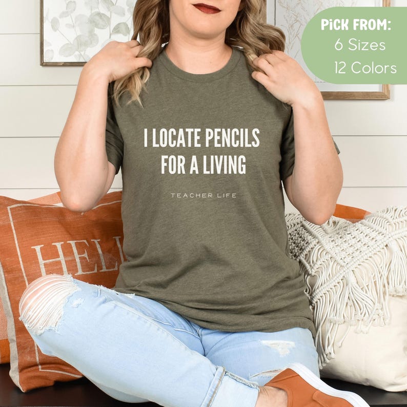 May include: A green t-shirt with the text "I locate pencils for a living" and "Teacher Life" in white lettering. The model is wearing the t-shirt and blue jeans.