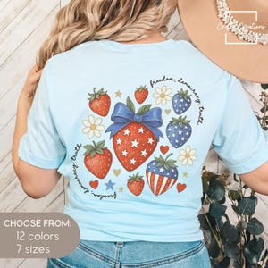 May include: Light blue t-shirt featuring a patriotic strawberry design with red, white, and blue elements. The design includes strawberries, stars, and the text "freedom, democracy, truth." The shirt is available in 12 colors and 7 sizes.