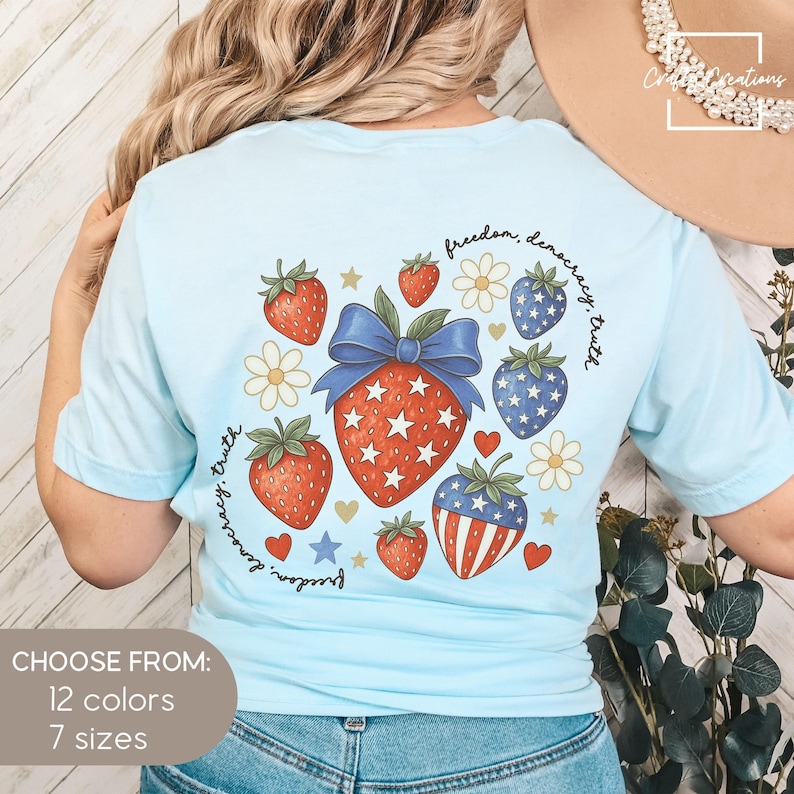 May include: Light blue t-shirt featuring a patriotic strawberry design with red, white, and blue elements. The design includes strawberries, stars, and the text "freedom, democracy, truth." The shirt is available in 12 colors and 7 sizes.