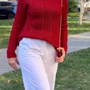 Puede incluir: Un suéter rojo vibrante de manga larga con cuello alto y un patrón de punto trenzado vertical, combinado con pantalones blancos. Un pequeño bolso de mano rojo con una fina cadena dorada se sostiene al costado, completando el conjunto.