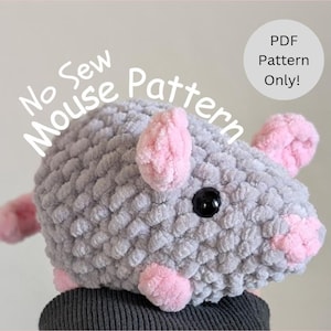 Puede incluir: Un ratón de crochet gris con orejas y nariz rosas. El ratón es un patrón sin costura, disponible para descargar en PDF. El texto "No Sew Mouse Pattern" está escrito encima del ratón.