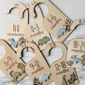 May include: Set of wooden hangers with dinosaur designs and numbers indicating age ranges for baby clothes. The hangers are labeled with the following age ranges: NB (newborn), 0-3 months, 3-6 months, 6-9 months, 9-12 months, 12-18 months, and 18-24 months.
