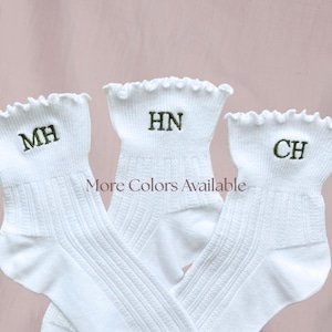 May include: Three white ribbed ankle socks with ruffled cuffs, each featuring custom green embroidered initials MH, HN, and CH. The text More Colors Available is centered between the socks. The socks have a vertical ribbed texture throughout.