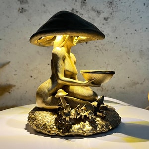 May include: A gold-coloured sculpture of a seated woman holding a bowl, under a mushroom-shaped canopy. The sculpture features naturalistic details, including small mushrooms and foliage at the base. The design has a whimsical, fantasy theme.