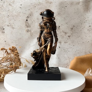 May include: A bronze-colored sculpture of a figure wearing an astronaut helmet and flowing gown, standing on a black base. The figure is posed as if walking, with one foot forward. The sculpture is on a white surface.
