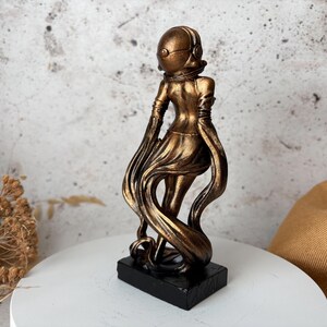 May include: A bronze-colored sculpture of a figure in a helmet and flowing garment, mounted on a black rectangular base. The figure's pose suggests movement, with the fabric trailing behind. The sculpture is set against a neutral background.