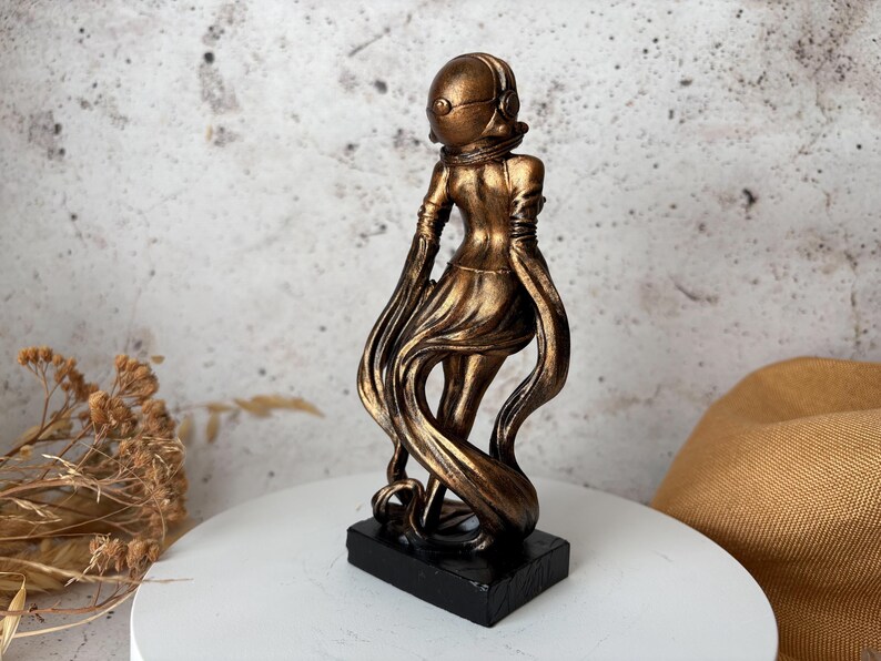 May include: A bronze-colored sculpture of a figure in a helmet and flowing garment, mounted on a black rectangular base. The figure's pose suggests movement, with the fabric trailing behind. The sculpture is set against a neutral background.