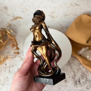 May include: A bronze-colored sculpture of a figure wearing an astronaut helmet. The figure is posed with flowing, stylized drapery. The sculpture is mounted on a black rectangular base. The figure is held in a hand.