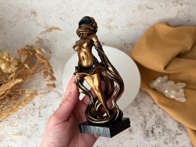May include: A bronze-colored sculpture of a figure wearing an astronaut helmet. The figure is posed with flowing, stylized drapery. The sculpture is mounted on a black rectangular base. The figure is held in a hand.