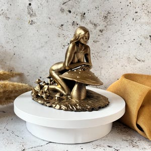 May include: A gold-colored figurine of a woman sitting on a mushroom. The figurine is detailed with a textured base and multiple mushrooms.
