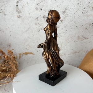 May include: A bronze-colored sculpture of a figure wearing a helmet and flowing gown, mounted on a black rectangular base. The statue has a futuristic aesthetic, with a detailed helmet and draped fabric.