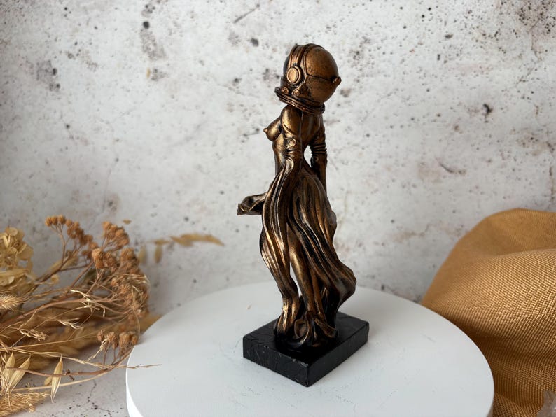May include: A bronze-colored sculpture of a figure wearing a helmet and flowing gown, mounted on a black rectangular base. The statue has a futuristic aesthetic, with a detailed helmet and draped fabric.