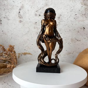 May include: A bronze-colored sculpture of a female figure with a helmet-like head covering and flowing drapery. The statue is mounted on a black rectangular base and stands on a white surface. The figure's form is stylized and elegant.